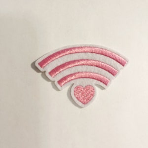 May include: A white fabric patch shaped like a Wi-Fi signal icon with three curved pink metallic stripes and a small pink heart at the base. The patch features a clean white border and a textured, shimmering finish on the pink elements.