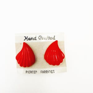 May include: A pair of bright red, shell-shaped pierced earrings. The earrings are displayed on a white card with the words "Hand Crafted" and "Pierced Earrings" printed on it. The earrings have a textured, ridged design.
