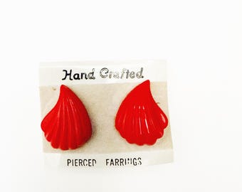 May include: A pair of bright red, shell-shaped pierced earrings. The earrings are displayed on a white card with the words "Hand Crafted" and "Pierced Earrings" printed on it. The earrings have a textured, ridged design.