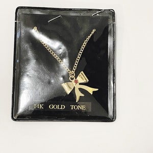 May include: A gold tone necklace with a bow pendant featuring a red gemstone. The necklace is packaged in a black plastic display card with the text "14K GOLD TONE".