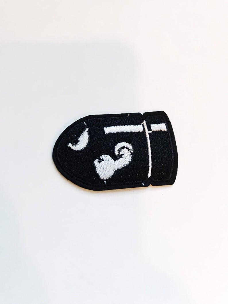 Puede incluir: Parche bordado negro con forma de bala con detalles blancos. El parche presenta una luna creciente blanca y un coraz&oacute;n blanco.