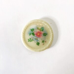 May include: A white ceramic button with a floral design. The button features a pink flower with white and blue flowers surrounding it. The button has a rope-like border.