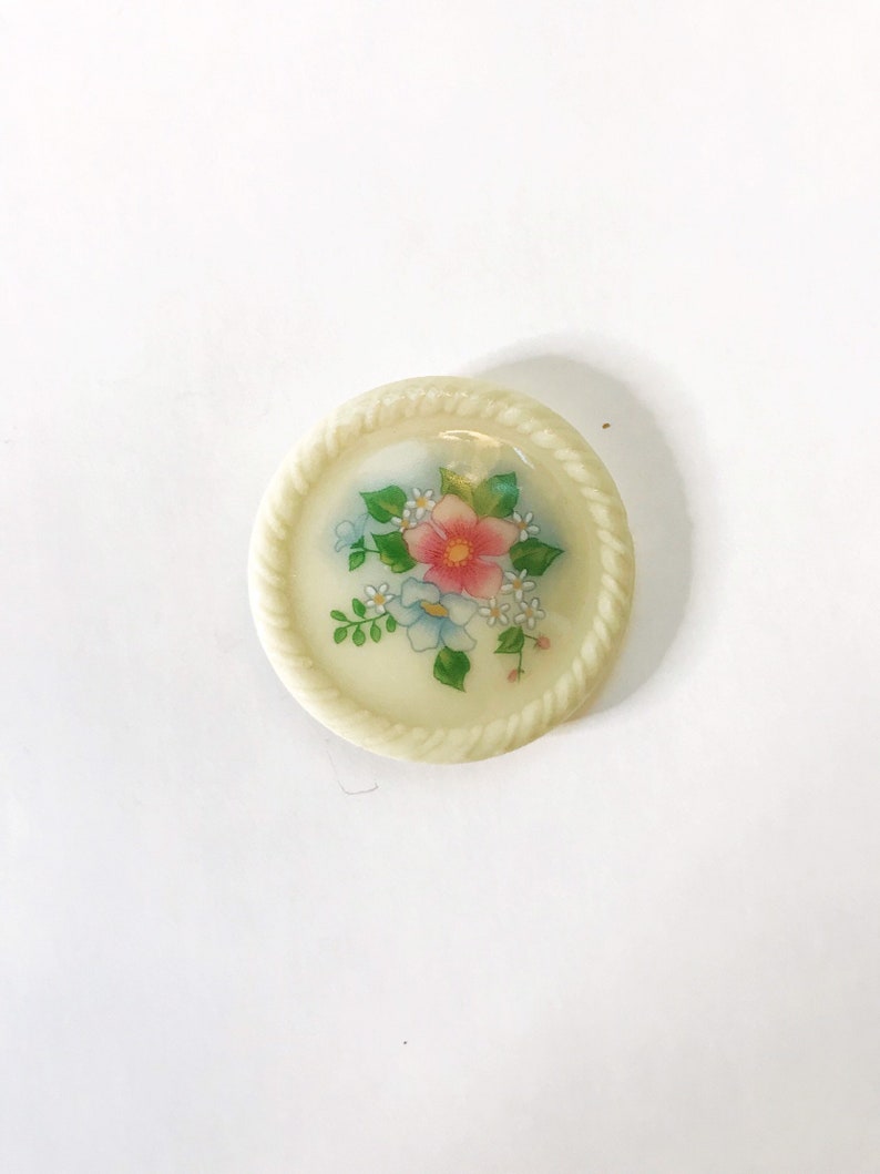 May include: A white ceramic button with a floral design. The button features a pink flower with white and blue flowers surrounding it. The button has a rope-like border.