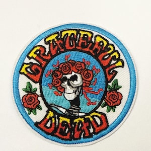 Puede incluir: Parche bordado circular con las palabras "Grateful Dead" que rodean una calavera adornada con rosas rojas. El texto es rojo y amarillo, sobre un fondo azul y blanco. Dos rosas rojas están a los lados del diseño.