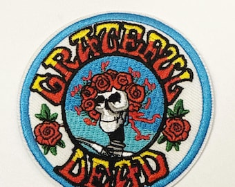 May include: Embroidered patch featuring a skull with a crown of red roses, surrounded by the words "Grateful Dead" in a circular design. The text is in red and yellow, set against a blue and white background. Two additional red roses are on the sides.