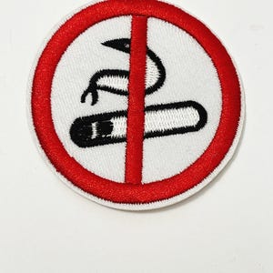May include: A white embroidered patch with a red circle and a black and white image of a cigarette with a line through it. The image is a no smoking symbol.