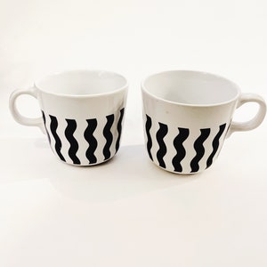 May include: Two white ceramic mugs with black wavy lines. The mugs have a handle on the right side.