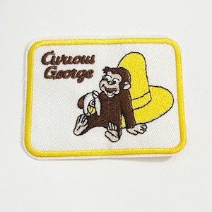 Puede incluir: Parche bordado con Curious George, un mono marrón, sosteniendo un plátano. El parche tiene un borde amarillo e incluye el texto "Curious George" en marrón. También se presenta un sombrero amarillo.