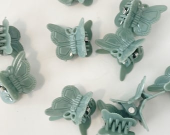 May include: A collection of small, sage green butterfly hair clips. Each clip features a detailed butterfly design with a spring-loaded clasp. The clips are scattered across a white surface, showcasing their delicate features.