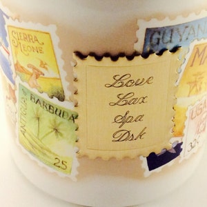 May include: A white ceramic mug with a gold-colored stamp design. The stamp features a gold background with the text "Love Lax Spa Dsk" written in cursive.
