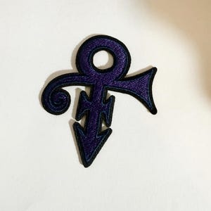 Peut inclure: Un écusson brodé représentant le symbole violet emblématique de Prince. Le motif se compose d'un sommet circulaire, d'une structure centrale en forme de croix, d'une base pointue et d'un élément latéral incurvé, le tout bordé de noir.