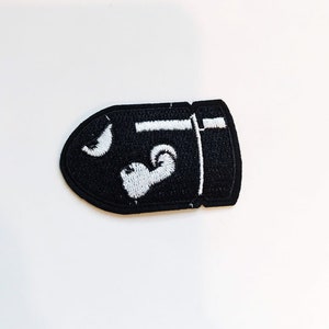 Puede incluir: Parche bordado negro con forma de bala con un contorno blanco y un dise&ntilde;o blanco en el interior.