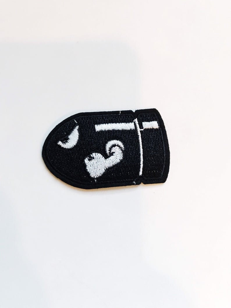Puede incluir: Parche bordado negro con forma de bala con un contorno blanco y un dise&ntilde;o blanco en el interior.