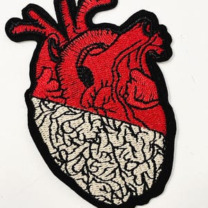 May include: An embroidered patch in the shape of an anatomical heart. The upper portion is stitched in vibrant red thread with black detailing, while the lower portion features a cream-colored brain-like pattern, all outlined in a thick black border.