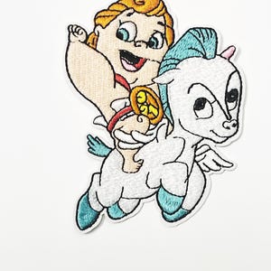 Könnte beinhalten: Ein gestickter Aufnäher mit einem Cartoon-Cupido, der auf einem geflügelten Pferd reitet. Cupido trägt eine rot-weiße Tunika und hält einen goldenen Pfeil. Das Pferd ist weiß mit blauen Flügeln und einer blauen Mähne.