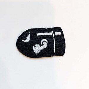 Puede incluir: Parche bordado negro con forma de bala con un contorno blanco y un dise&ntilde;o blanco en el interior.