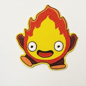 May include: Embroidered patch of a cartoon fire demon. The patch has a yellow body with red flames, a smiling face with large white eyes and a black mouth. It has small brown arms and feet. The patch is set against a white background.
