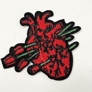 May include: An embroidered patch featuring an anatomical heart in vibrant red with black outlines and details. The heart is pierced by several green arrows, each tipped with red, creating a striking contrast against the red and black heart design.
