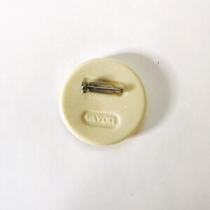 May include: A white round pin with a silver pin back. The pin has the word "Avon" embossed on the front.