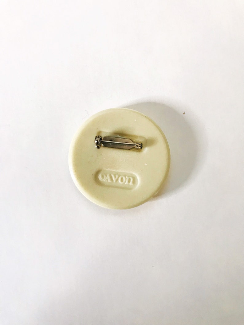 May include: A white round pin with a silver pin back. The pin has the word "Avon" embossed on the front.