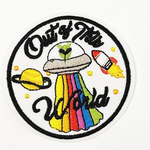 May include: Embroidered patch with a white background and a black border. The patch features a UFO with a rainbow trail, a yellow planet, and a red rocket. Text reads "Out of This World."