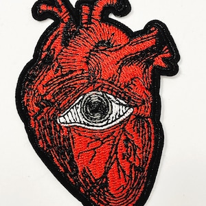 May include: Embroidered patch featuring a red anatomical heart with a black outline. The heart has a white eye in the center. The patch is a unique accessory for jackets, bags, or other fabric items.