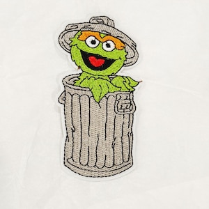 May include: Embroidered patch of Oscar the Grouch from Sesame Street, peeking out of a grey bin. Oscar is green with orange eyebrows and a red mouth, wearing a grey hat. The patch is set against a white backdrop.