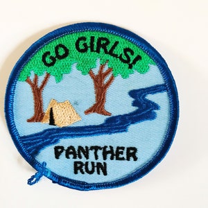 May include: A blue and green embroidered patch with the text "GO GIRLS!" and "PANTHER RUN". The patch features a tent, trees, and a river.