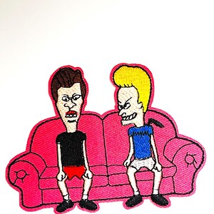 Op de afbeelding: Een geborduurde patch met twee cartoonfiguren, een met bruin haar en de andere met blond haar, zittend op een roze bank. Beide figuren zijn casual gekleed en hebben boze gezichtsuitdrukkingen.