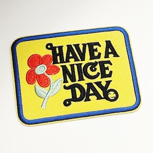 May include: Yellow embroidered patch with a blue border. The patch features a red flower with a green stem and the text "HAVE A NICE DAY" in black with a smiley face.