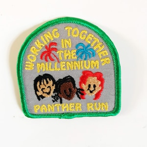 Op de afbeelding: Een geborduurde patch in groen en grijs met de tekst "Working Together in the Millennium" en "Panther Run". De patch toont drie cartoon gezichten van mensen met verschillende huidtinten.