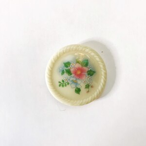 May include: A white ceramic button with a floral design. The button features a pink flower with blue and white flowers surrounding it. The button has a rope-like border.