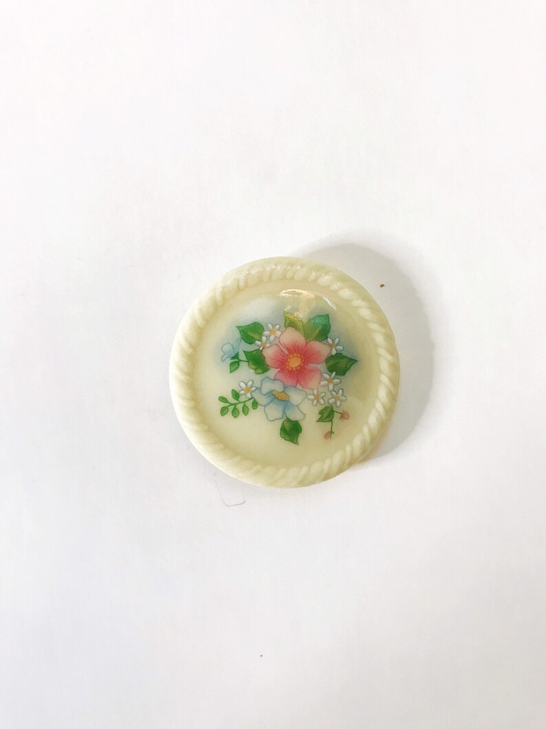 May include: A white ceramic button with a floral design. The button features a pink flower with blue and white flowers surrounding it. The button has a rope-like border.