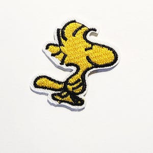 以下が含まれることがあります： 黄色い漫画の鳥のキャラクター、ウッドストックをあしらった刺繍入りのアイロン接着ワッペン。黒い糸で縁取りされ、白い枠が付いたデザインで、頭を右側に向けた立ち姿の鳥が描かれています。
