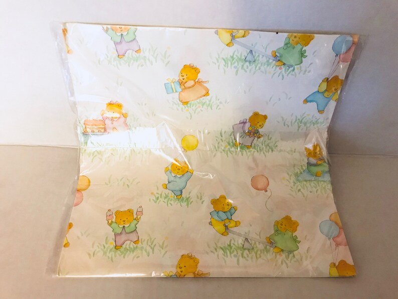 May include: A sheet of wrapping paper with a light yellow background and a repeating pattern of teddy bears wearing different colored clothes and holding balloons, presents, and other party favors.