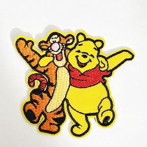 May include: Embroidered patch featuring Winnie the Pooh and Tigger. Winnie the Pooh is yellow and wearing a red shirt. Tigger is orange and black with a red tail. Both characters are smiling and have their arms outstretched.