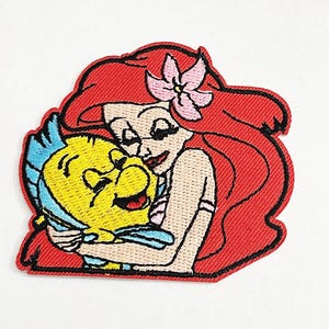 Op de afbeelding: Geborduurde patch met Ariel uit De Kleine Zeemeermin die Botje omhelst. Ariel heeft rood haar en een roze bloem. Botje is geel met blauwe vinnen. De patch heeft een rode rand op een witte achtergrond.