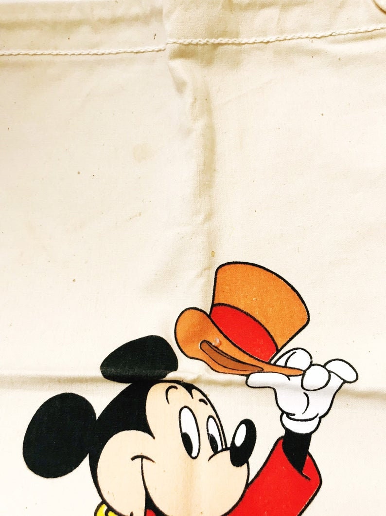 Może przedstawiać: Kremowa torba typu tote z kresk&oacute;wkową ilustracją Myszki Miki. Miki, w czerwonej kurtce, czarnych uszach i białych rękawicach, trzyma brązowo-czerwony kapelusz. Torba ma prosty design z przeszyciem.