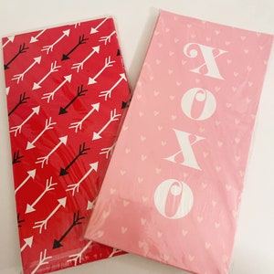 Puede incluir: Dos rollos de papel de regalo para el Día de San Valentín. Un rollo tiene un fondo rojo con flechas negras y blancas. El otro rollo tiene un fondo rosa con corazones blancos y la palabra "XOXO" en blanco.