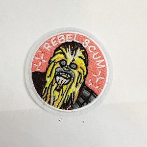 May include: A round patch featuring a Wookiee character with yellow fur and a brown face. The patch has the text "LY REBEL SCUM" in white letters on a red background, surrounded by a white border.