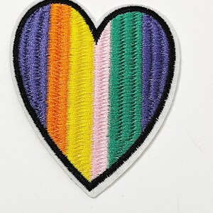 May include: Embroidered heart-shaped patch with a rainbow design. The patch features vertical stripes in purple, orange, yellow, pink, green, and blue. The heart is outlined in black and has a white border.