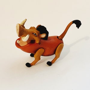 Könnte beinhalten: Eine braune und schwarze Plastikfigur von Pumbaa aus Disneys Der König der Löwen. Die Figur hat eine große Schnauze mit weißen Stoßzähnen und eine schwarze Mähne. Die Figur ist in einer laufenden Pose mit erhobenem Schwanz.