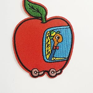 May include: Embroidered patch featuring a red apple with a green leaf and a black outline. Inside the apple is a blue window with a cartoon worm wearing a bow tie. The apple has wheels, suggesting a vehicle.