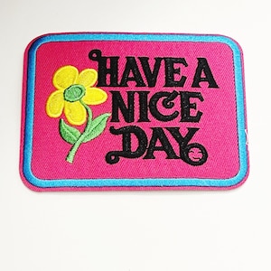 May include: Pink fabric patch with a blue border. The patch features a yellow flower with a green stem and the text "HAVE A NICE DAY" in black.