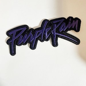 Puede incluir: Un parche bordado que presenta el texto Purple Rain en una fuente de escritura morada estilizada. Las letras están delineadas con un borde negro grueso, creando un contraste marcado con el hilo morado. El diseño se presenta como una pieza.