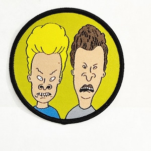 Op de afbeelding: Een ronde patch met twee stripfiguren met overdreven gezichtsuitdrukkingen. De ene heeft blond haar, de andere bruin haar. De patch heeft een gele achtergrond met een zwarte rand.