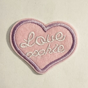 May include: A heart-shaped patch made of light pink felt with a purple embroidered border. The center features the white embroidered cursive text "Love Rosie" stitched in a flowing script style across the middle of the pink heart surface.