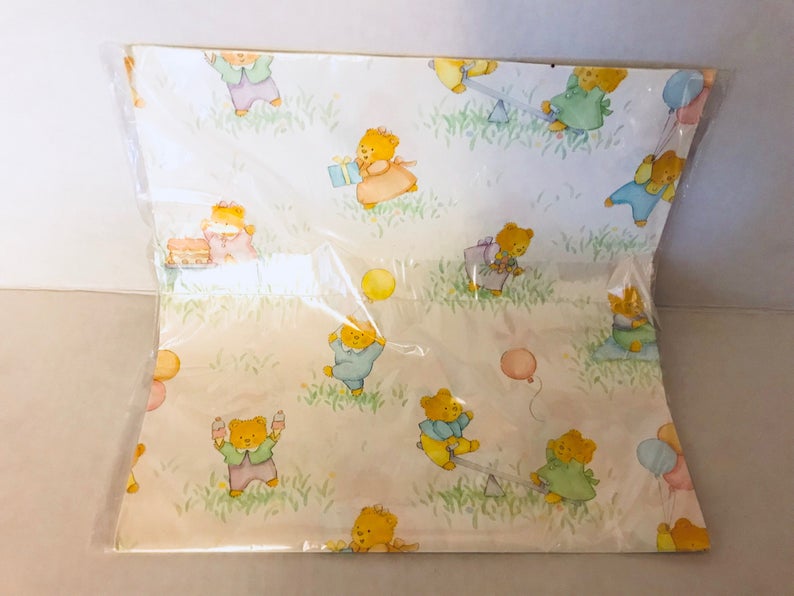 May include: A sheet of wrapping paper with a light yellow background and a repeating pattern of teddy bears wearing different colored clothes and holding balloons, presents, and other party favors.