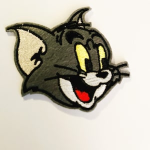 May include: A gray embroidered patch of Tom the cat from the cartoon Tom and Jerry. Tom is smiling with his mouth open and his tongue sticking out. He has yellow eyes and a white nose.