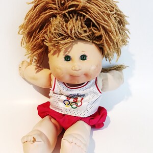 May include: A Cabbage Patch Kid doll with blonde yarn hair wearing a white tank top with the Olympic logo and a red skirt.